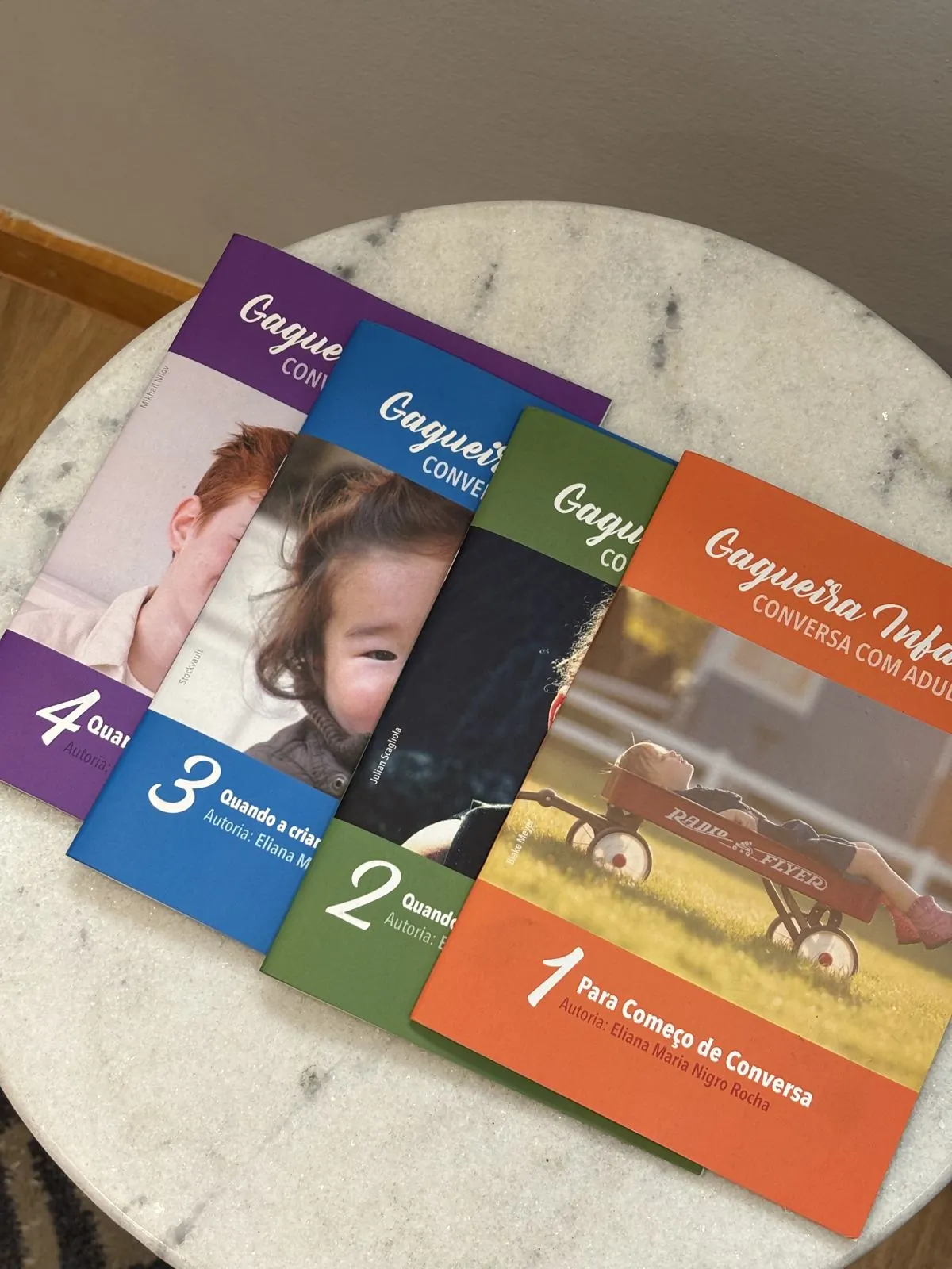 Série de folhetos Gagueira Infantil: conversa com adultos, volumes 1 a 4, sobre uma mesa de mármore